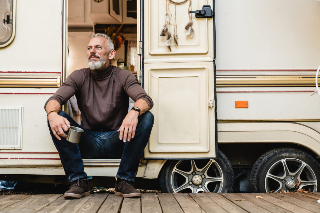 senior man with a beard sitting on the doorstep of the motor home holding iron mug